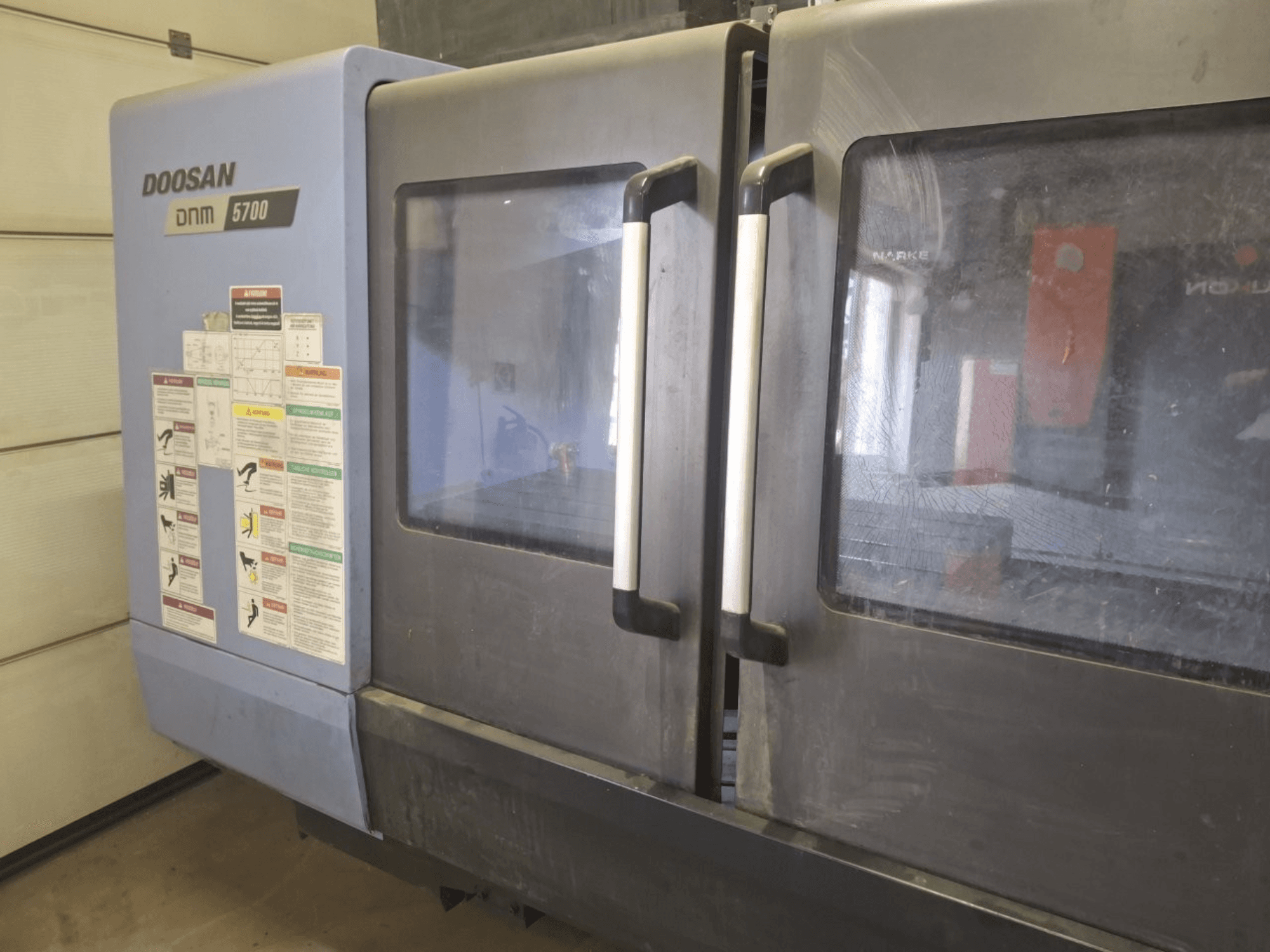 Prednji pogled na vertikalni obradni centar DOOSAN DNM 5700, s dvostrukim vratima i upravljačkim pločama s uputama za uporabu.