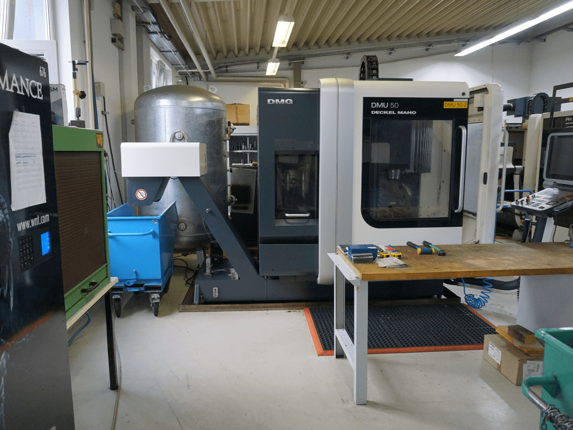 Glodalica DMG DMU 50 u radionici, prednji pogled, s alatima na drvenom stolu i plavom kantom na kotačima u blizini.