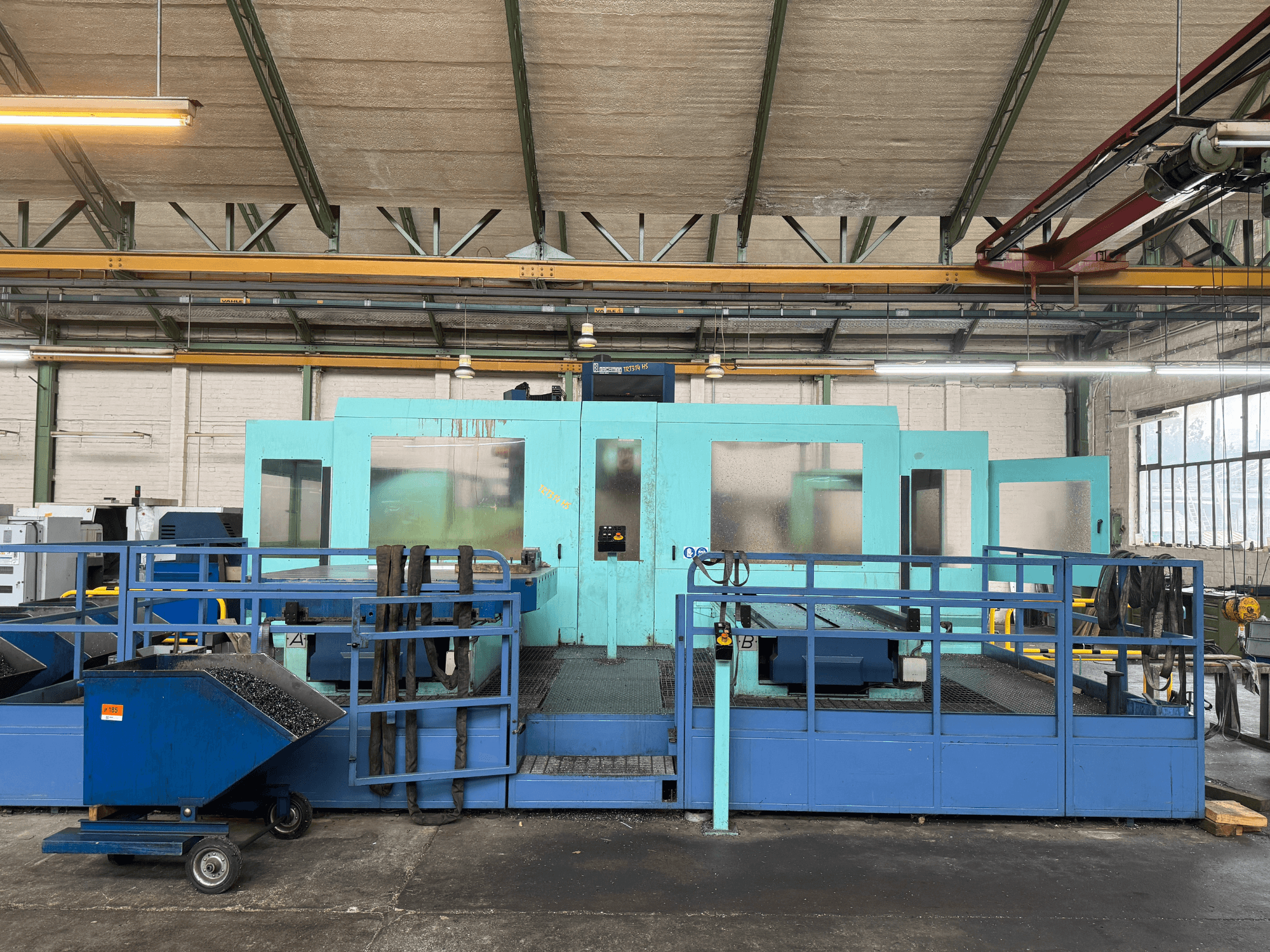 Industrijski plavi CNC stroj sa zaštitnim pločama, prednji pogled, s prostorom za pohranu alata i kolicima za sakupljanje otpada sa strane.