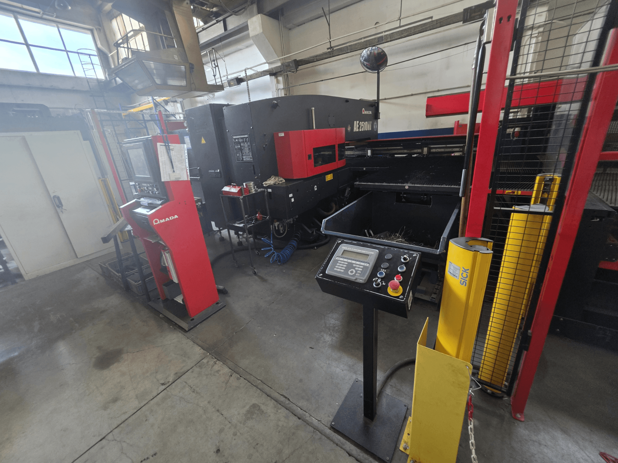CNC stroj za probijanje AMADA AE-2510NT, prednji pogled s upravljačkom pločom, sigurnosnim značajkama i okolnim industrijskim okruženjem.