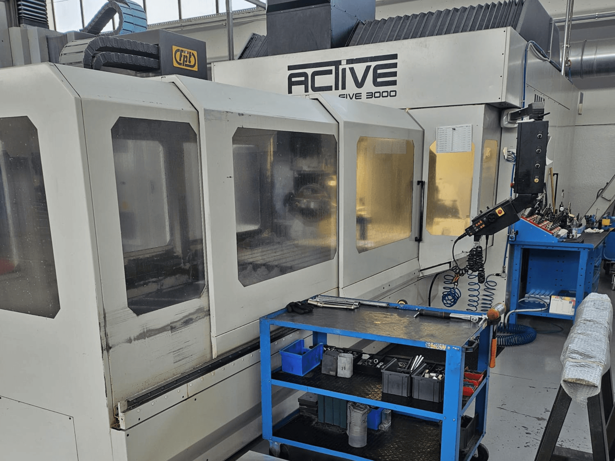 Aktivni CNC stroj 3000 u radionici, prednji pogled s alatima na plavim kolicima, vidljive kontrole i radni prostor unutra.