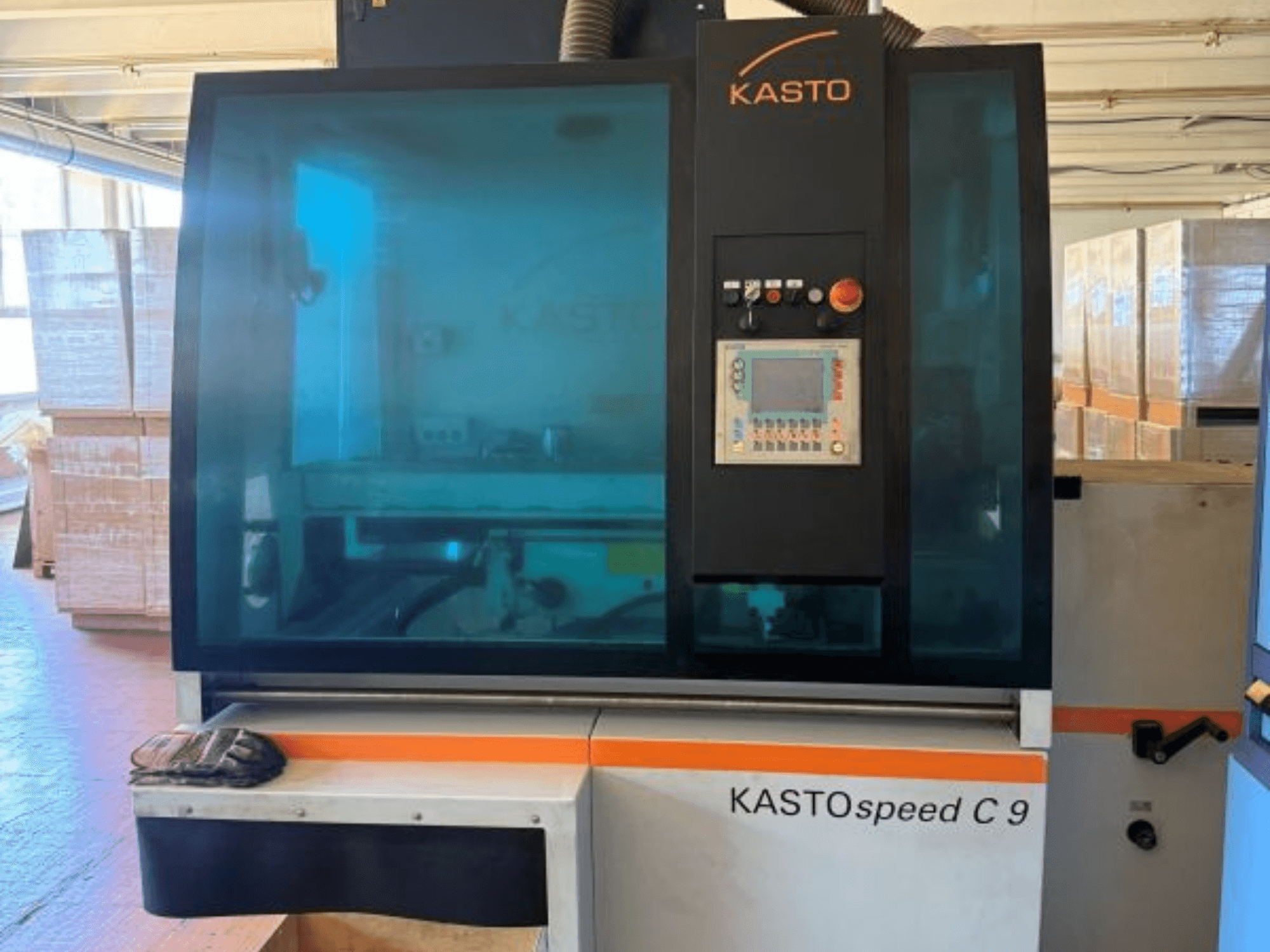 Prednji pogled na kružnu pilu za metal KASTO KASTOspeed C 9, s upravljačkom pločom i sigurnosnim staklom, s drvenim sanducima u pozadini.