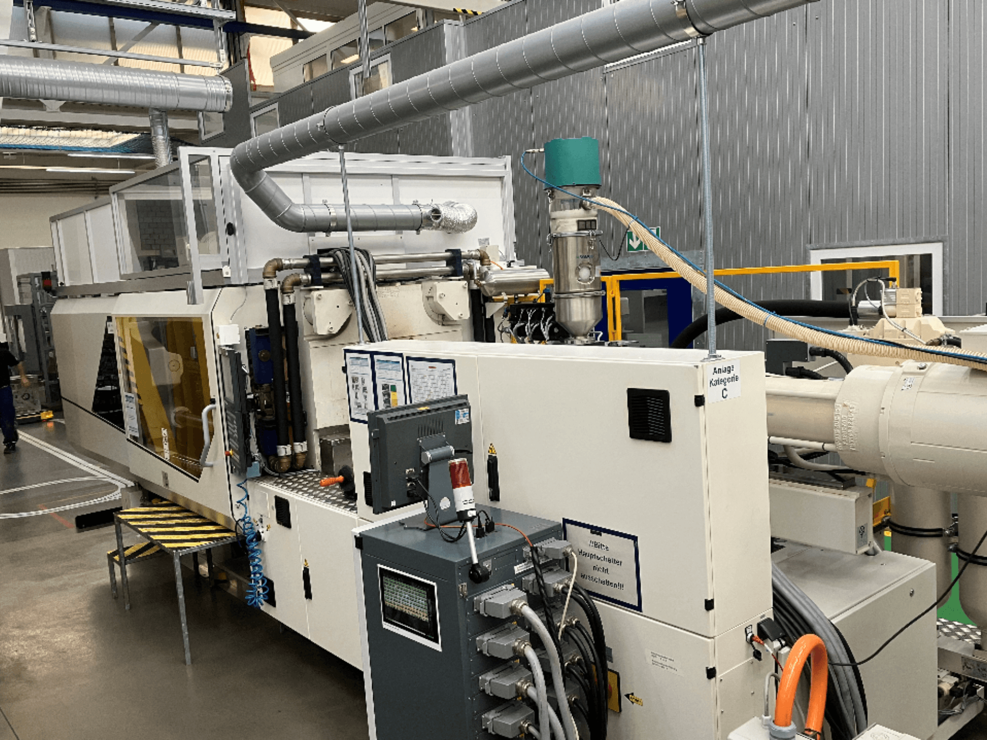 Stroj za brizganje plastike, prednji pogled, s naljepnicom robne marke, upravljačkom pločom i vidljivim crijevima u tvorničkom okruženju.