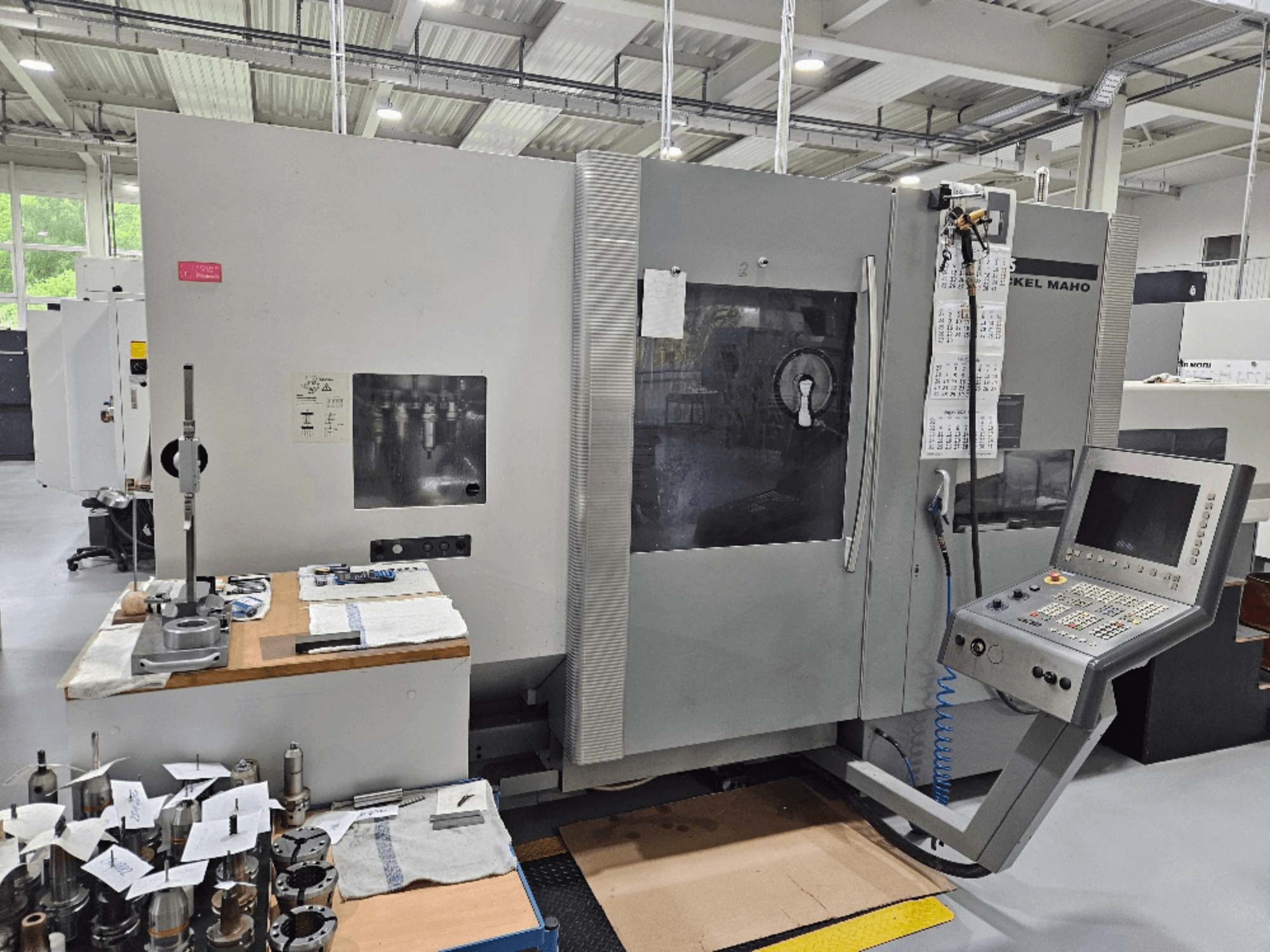 Prednji pogled na stroj DMG Mori DMU 70 s upravljačkom pločom, alatima na drvenom stolu i raznim radnim komadima u blizini.