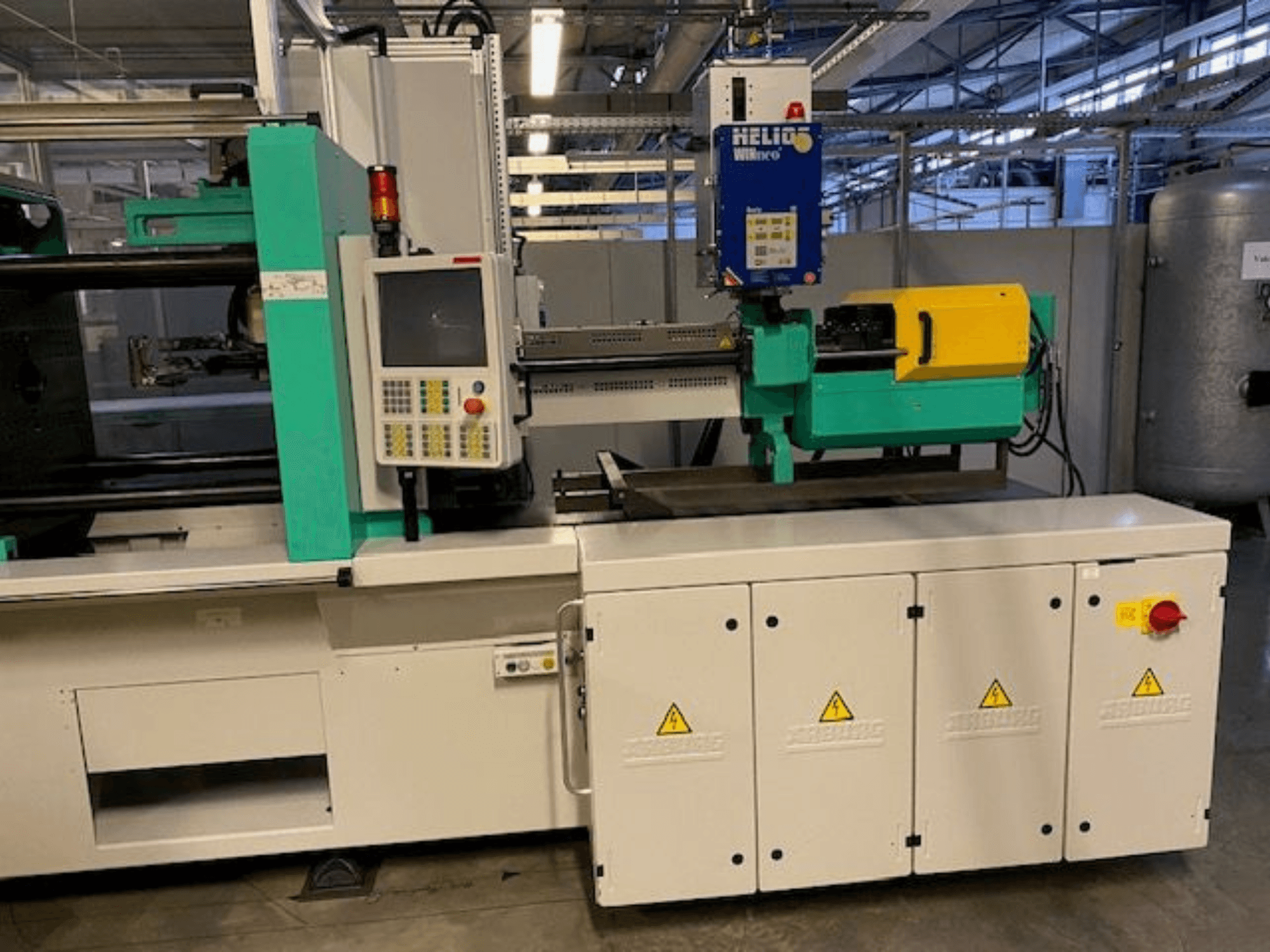 Industrijski stroj za brizganje plastike, model HELIO, prednji pogled, s upravljačkom pločom, sigurnosnim štitnicima i tipkom za zaustavljanje u nuždi.