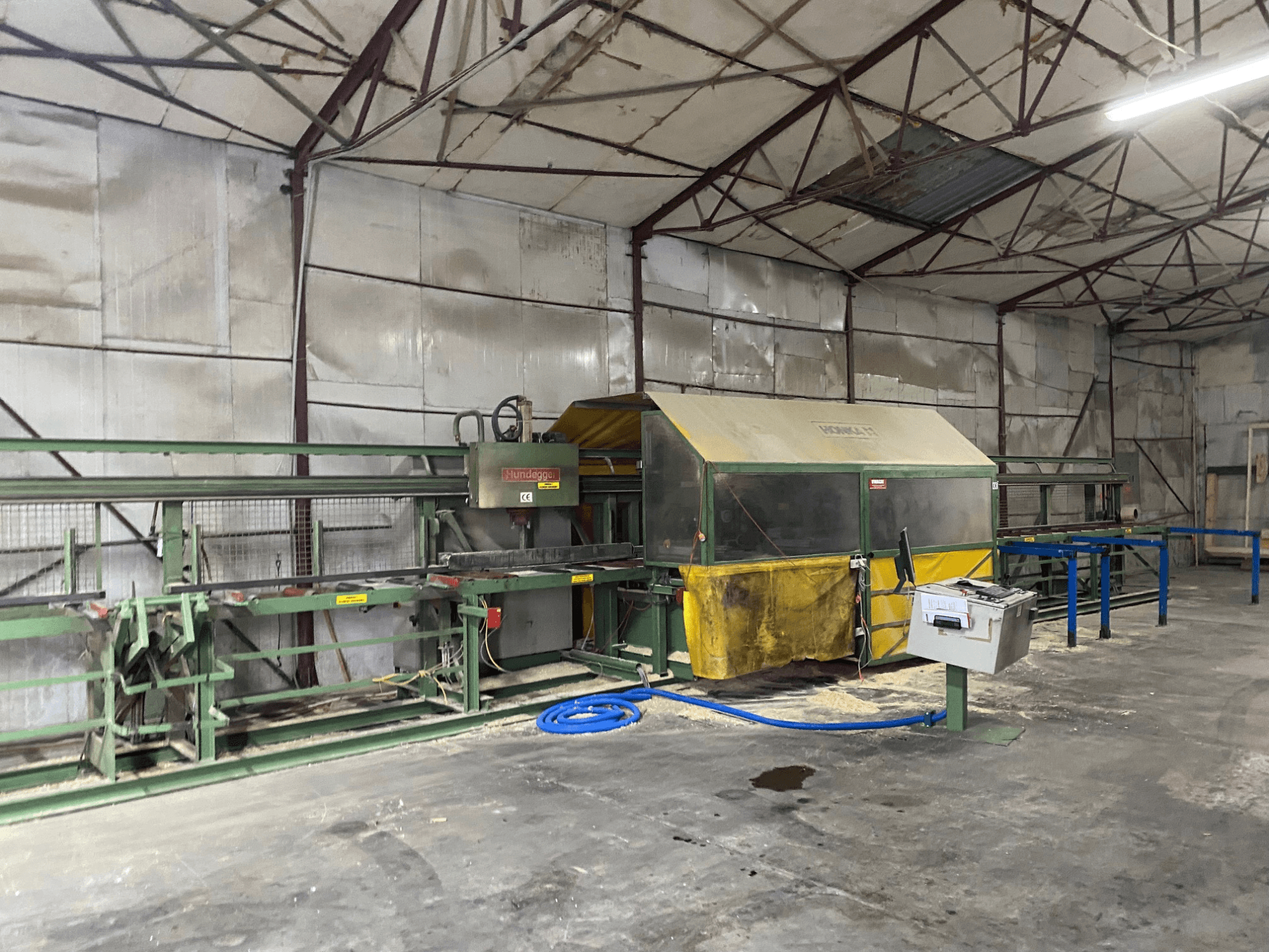 CNC centar za obradu drva HUNDEGGER K1 u radionici, bočni pogled koji prikazuje strojeve, upravljačke elemente i piljevinu na podu.