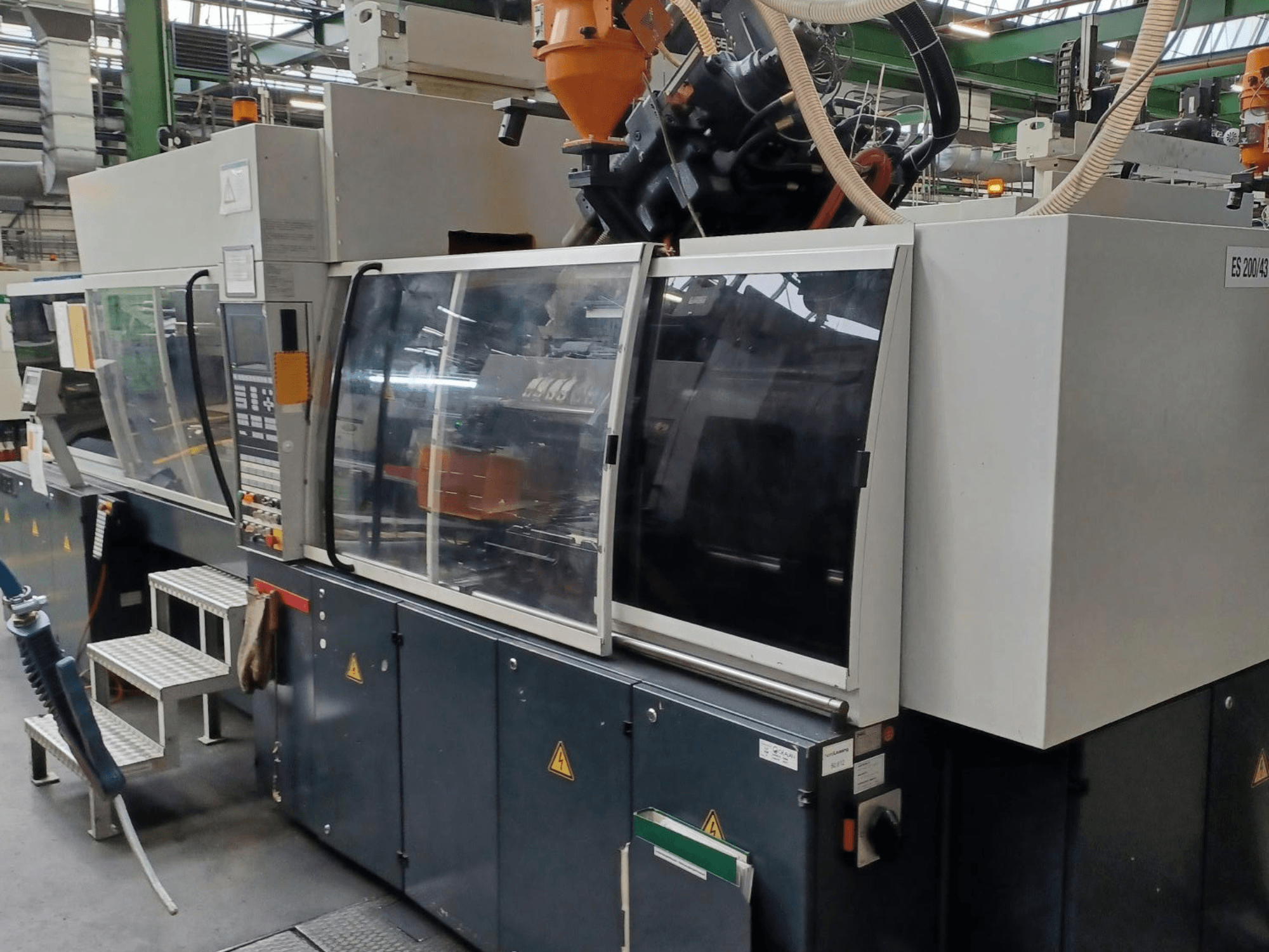 Prednji pogled na stroj za brizganje plastike, nepoznate marke i modela, s prozirnim sigurnosnim vratima i upravljačkom pločom.