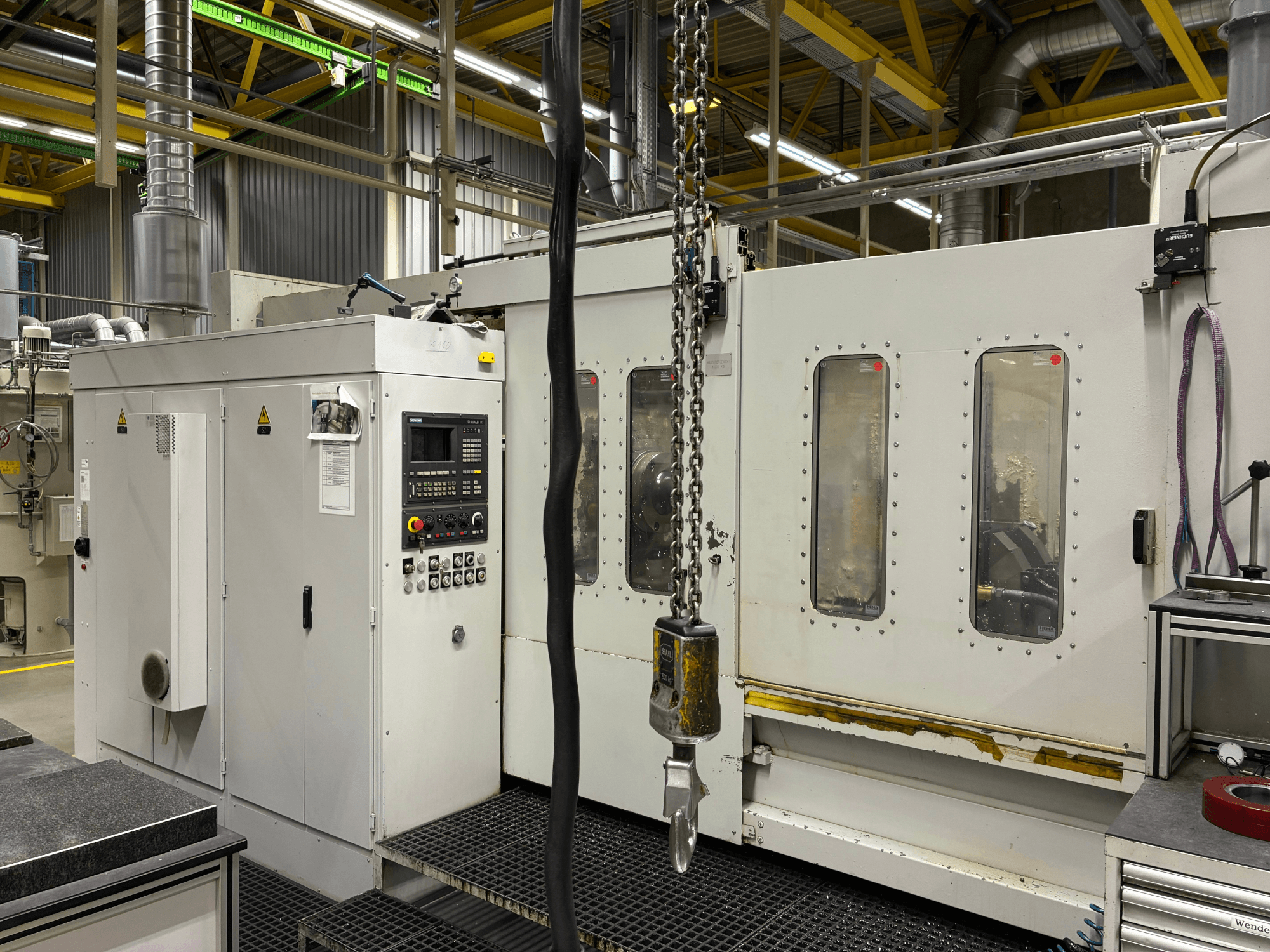 Industrijski CNC stroj s upravljačkom pločom, bočni pogled; ima prozore, lance i metalne komponente u tvorničkom okruženju.