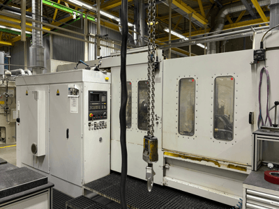 Industrijski CNC stroj s upravljačkom pločom, bočni pogled; ima prozore, lance i metalne komponente u tvorničkom okruženju.